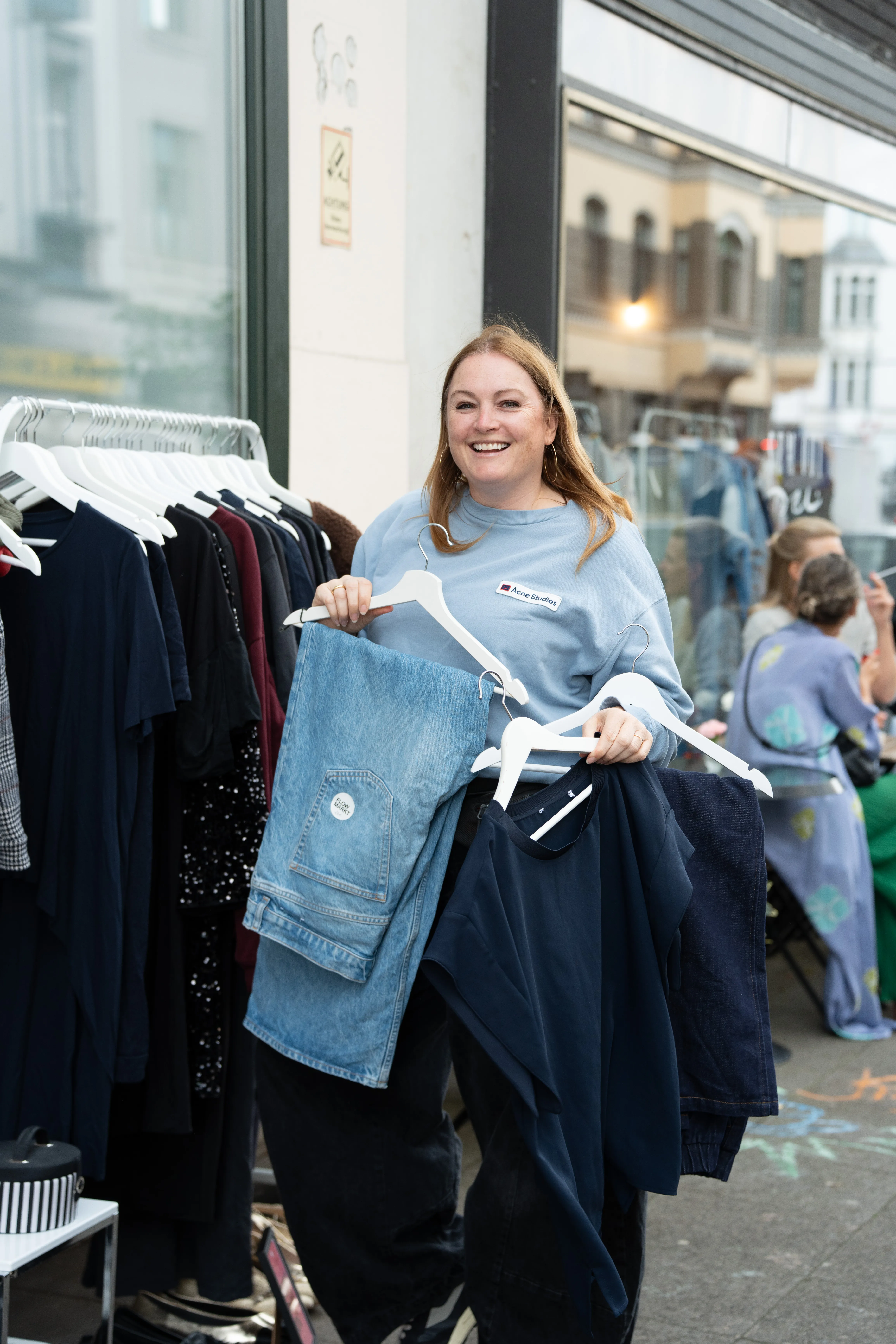 Lächelnde Besucherin an der Kleiderstange auf dem Flowmarkt, Fundstücke mit Flowmarkt-Anhänger in der Hand